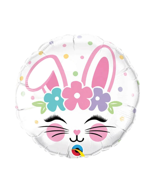 A round foil balloon shaped like a bunny face, with large pink bunny ears, a flower crown in pink, teal, purple, and green, closed eyes with long black lashes, pink cheeks, and small multicolored polka dots on a white background. A small colorful logo is visible at the bottom center.